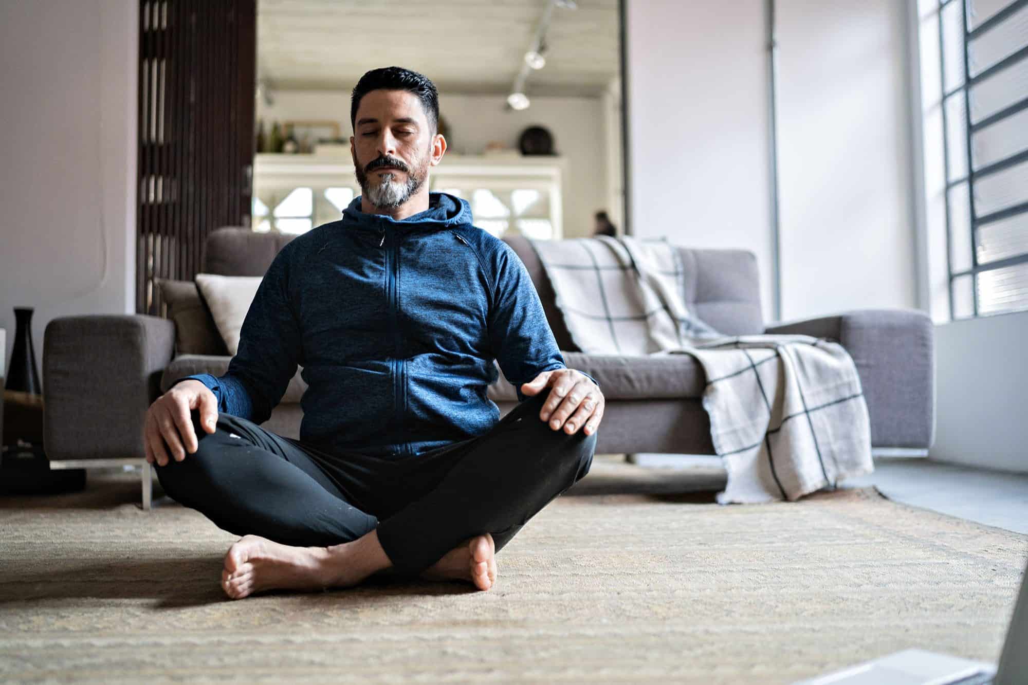 Man meditating in living room, sitting cross-legged on rug